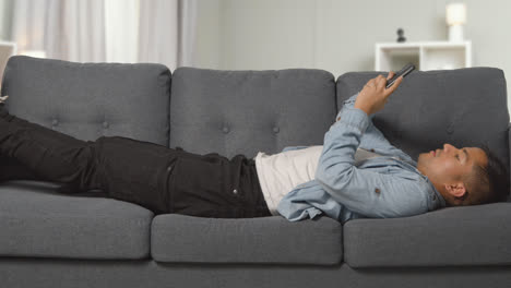 Young-Man-Lying-On-Sofa-At-Home-Scrolling-Through-Mobile-Phone